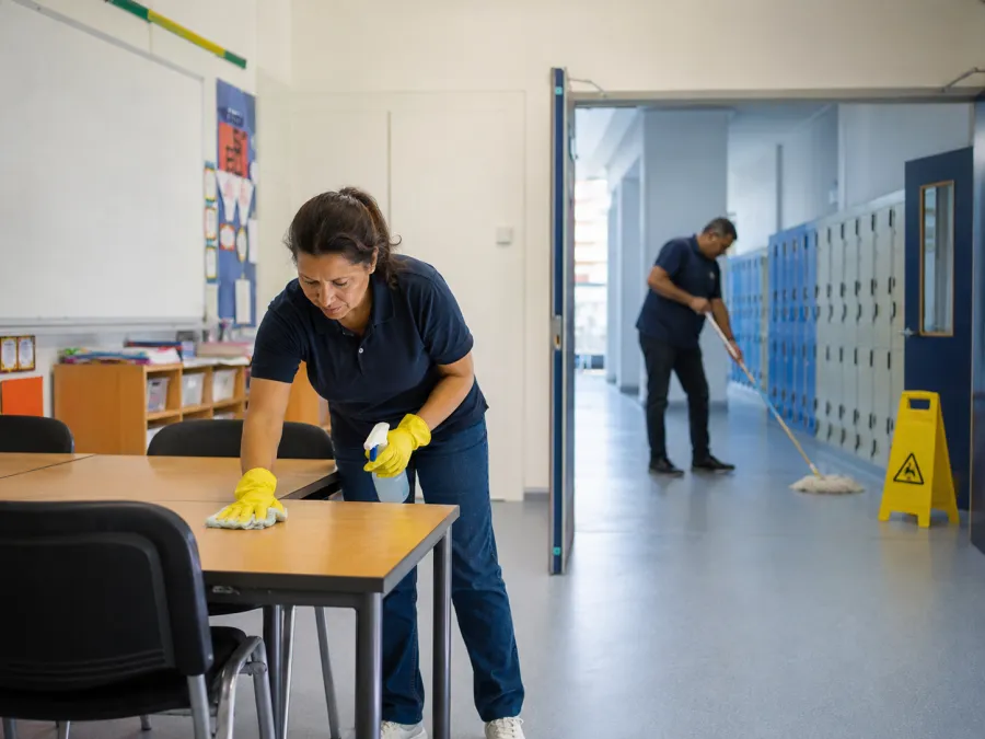 Cleaner wiping desks in a school classroom with lockers in the corridor beyond