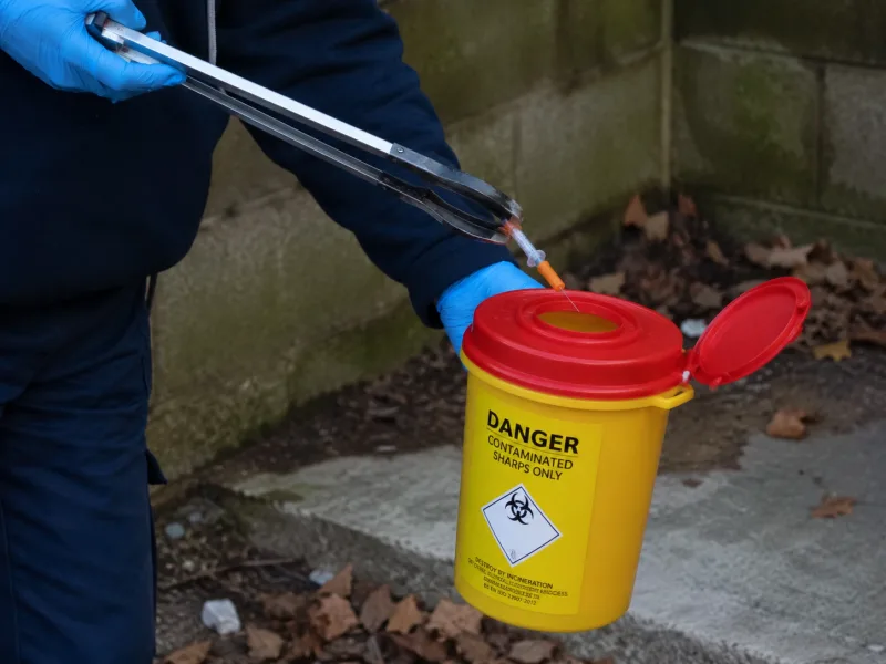 Gloved operative using a litter-picker to place a needle into a yellow sharps bin